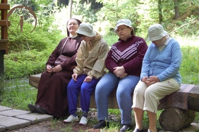 s. Szymona z Panią Anną, Panią Emilią i Panią Anną słuchają w pozycji siedzącej na ławce z pnia drzewa przy Kapliczce