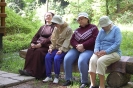 s. Szymona z Panią Anną, Panią Emilią i Panią Anną słuchają w pozycji siedzącej na ławce z pnia drzewa przy Kapliczce