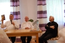 Przy prezydialnym stole siedzą ks. Mirosław prezentując książkę ks. Grzegorza oraz słuchający ks. Grzegorz i ks. Antoni
