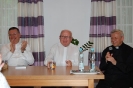Przy prezydialnym stole siedzą bijący brawo ks. Mirosław, uśmiechnięty ks. Grzegorz oraz radośnie mówiący ks. Antoni